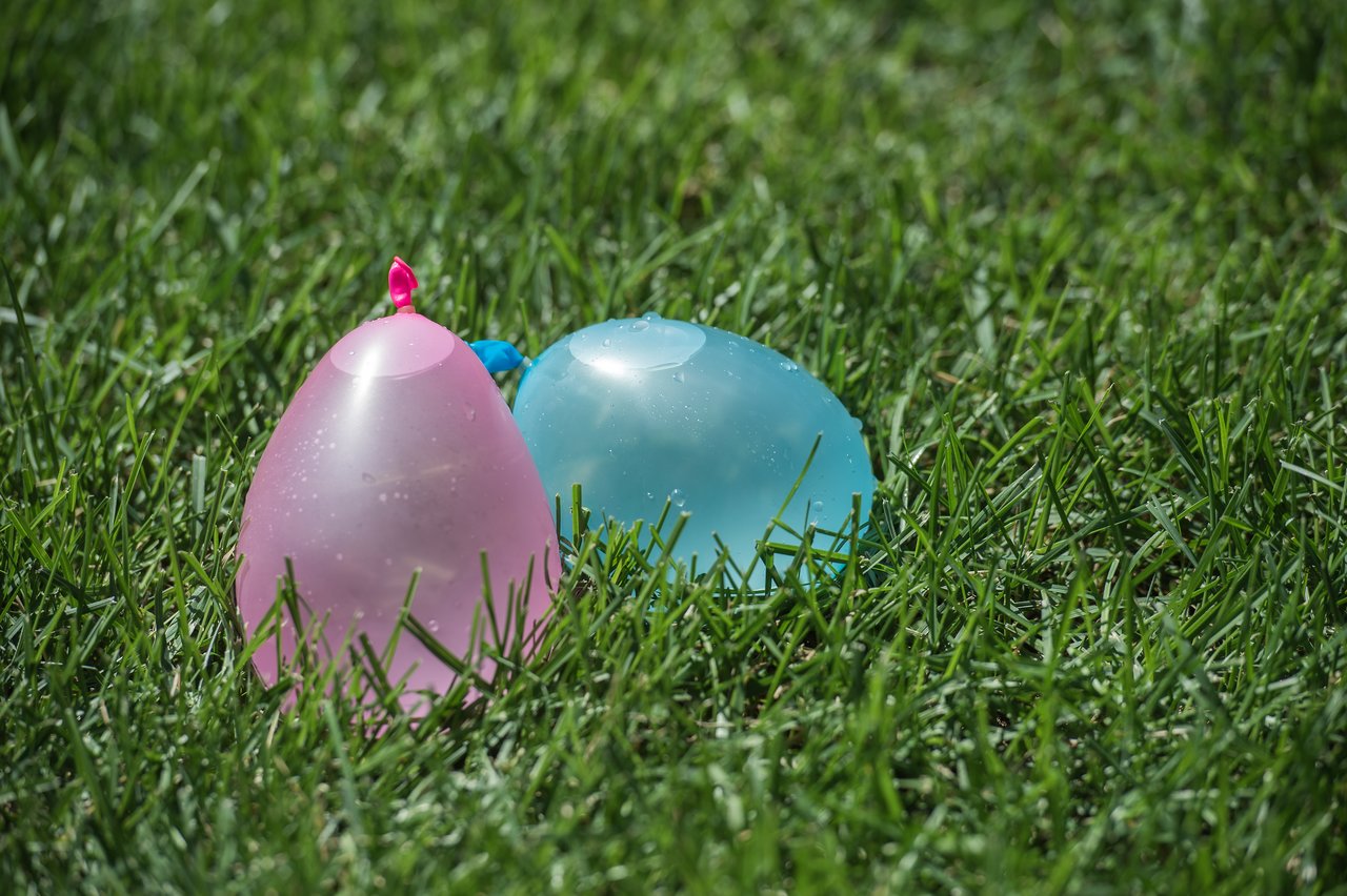 Two water balloons, one pink and one blue, laying on the grass, glistening with small droplets of water.