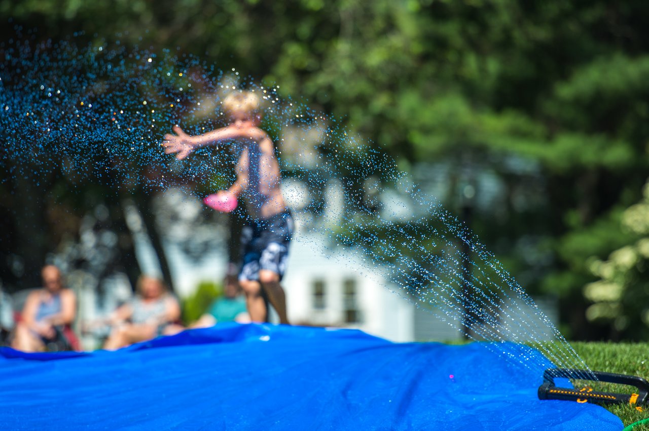 A child plays on a blue slip-and-slide, splashing water while preparing to throw a water balloon.