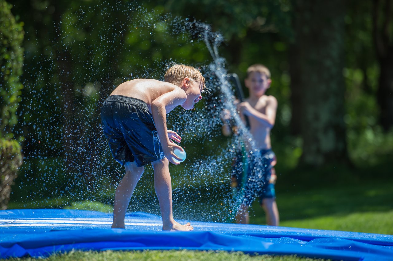 Two boys play with water outdoors; one sprays a hose while the other holds a water balloon on a slide.