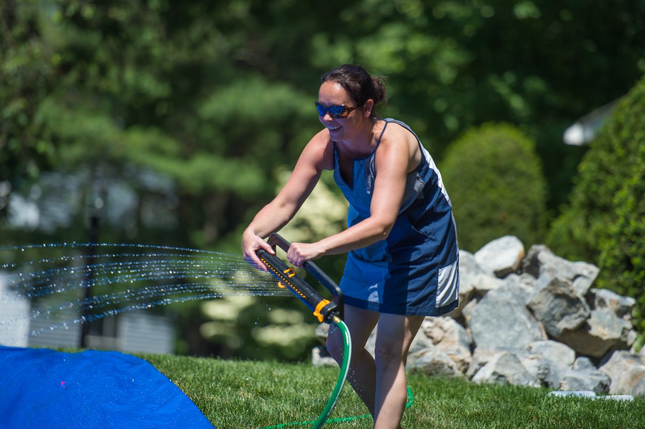 A woman in sunglasses sprays water from a hose onto a blue slip-and-slide.