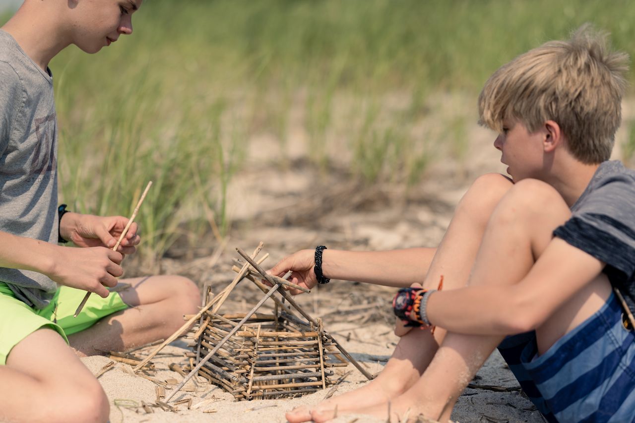 Two children sit on the sandy beach, building a small house structure out of sticks.