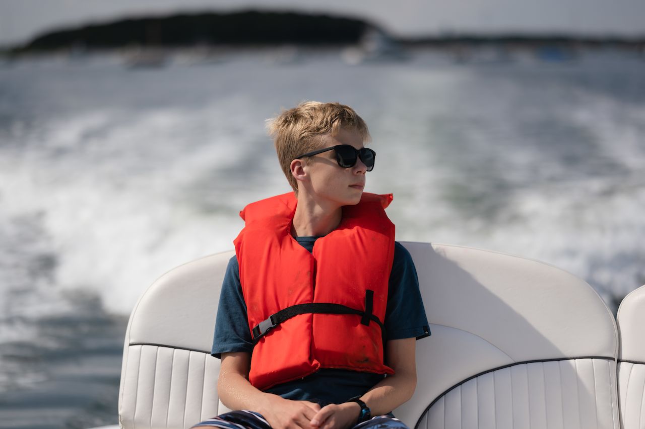 A person wearing a red life jacket and sunglasses sits on a moving boat, looking to the side.