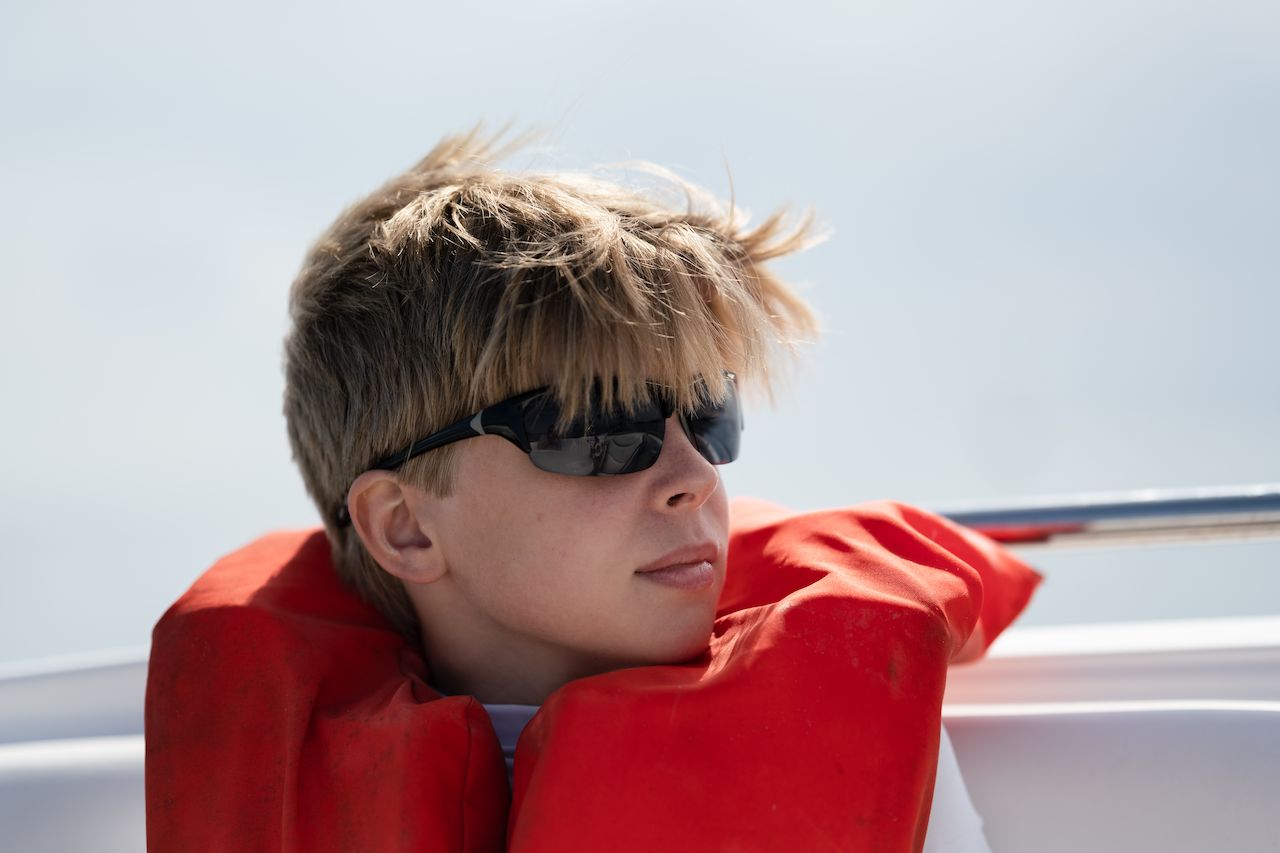 A person wearing sunglasses and a red life jacket sits on a boat, looking off into the distance.