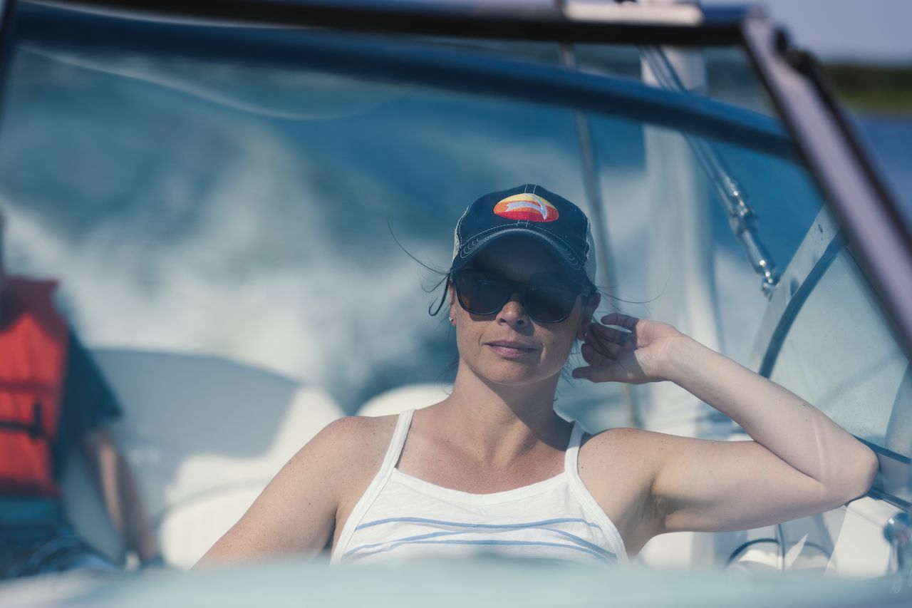 A woman wearing sunglasses and a cap relaxes on a moving boat, leaning back with one arm raised.