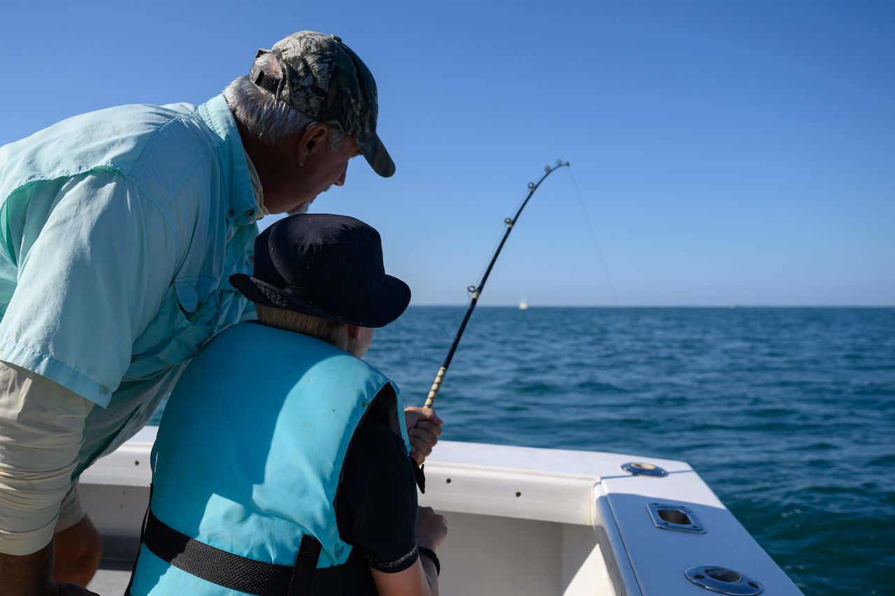 A child wearing a life vest holds a fishing rod while an older man watches on a boat in the ocean.