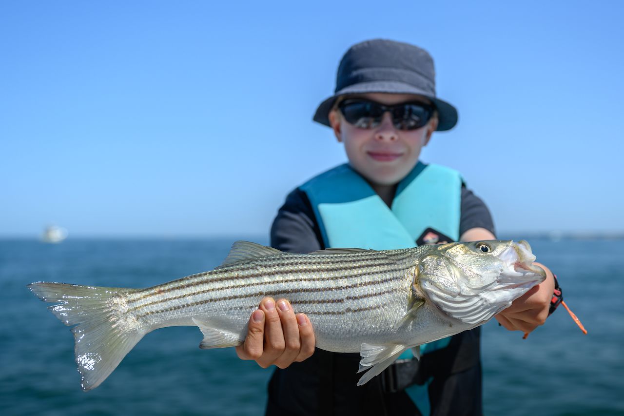A person wearing a hat and life vest holds a freshly caught striped fish while deep sea fishing.