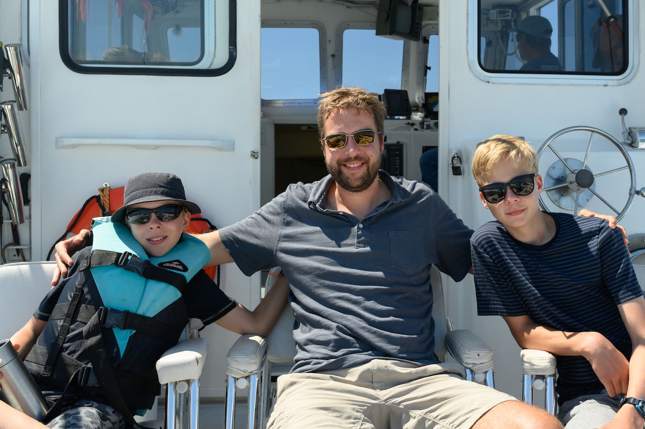 A man and two boys sit on a boat, smiling and wearing sunglasses, ready for deep sea fishing.