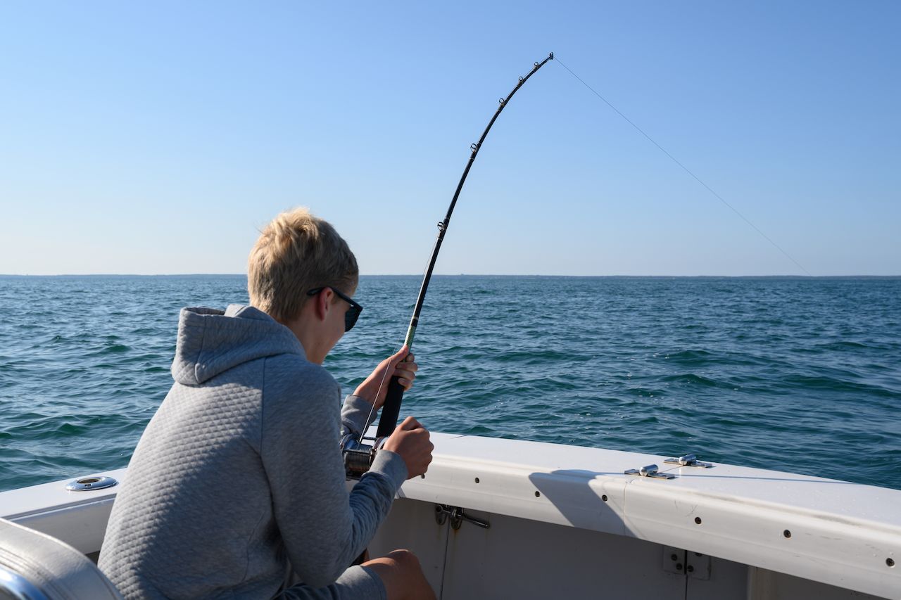 A person wearing sunglasses and a hoodie holds a fishing rod while deep sea fishing on a boat.