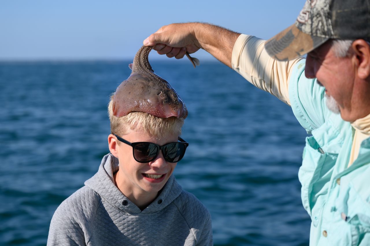 A man playfully places a small stingray on a boy's head while fishing in the ocean.