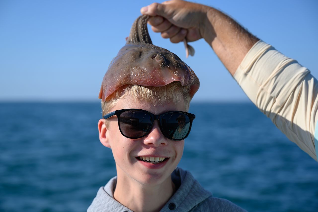 A person wearing sunglasses smiles as someone places a small stingray on their head during a fishing trip.
