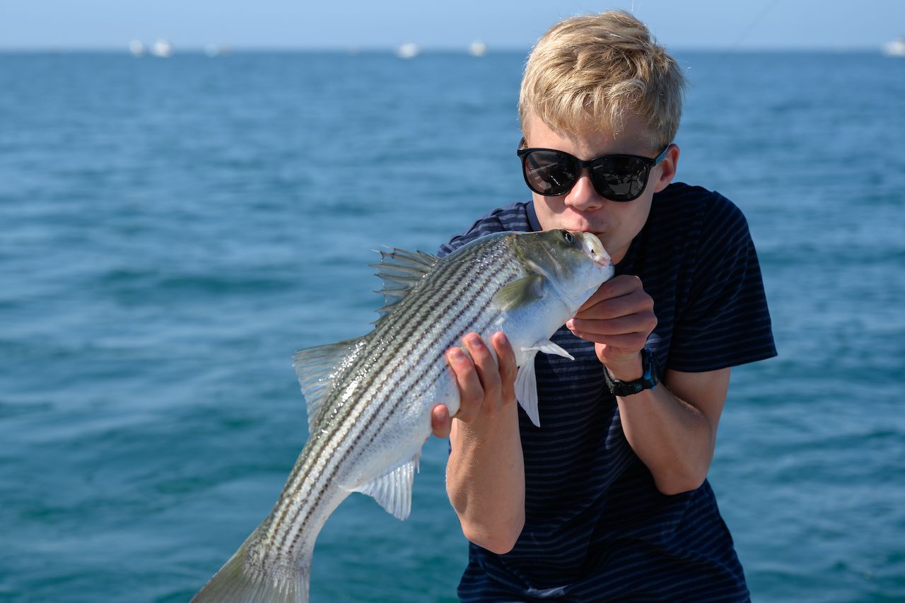 A person wearing sunglasses holds a freshly caught fish and playfully kisses it while on a boat in the ocean.
