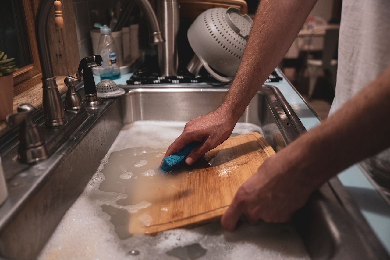 A person washes wooden cutting boards in a sink filled with soapy water, scrubbing with a blue sponge.