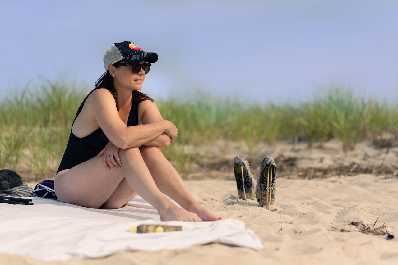 A woman in a swimsuit sits on a beach towel, drying off in the sun with shoes stuck in the sand.