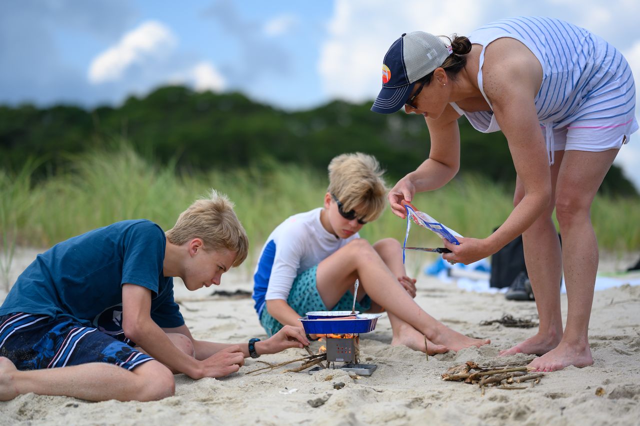 A woman and two children cook hot dogs on a small beach stove, gathering sticks to keep the fire going.