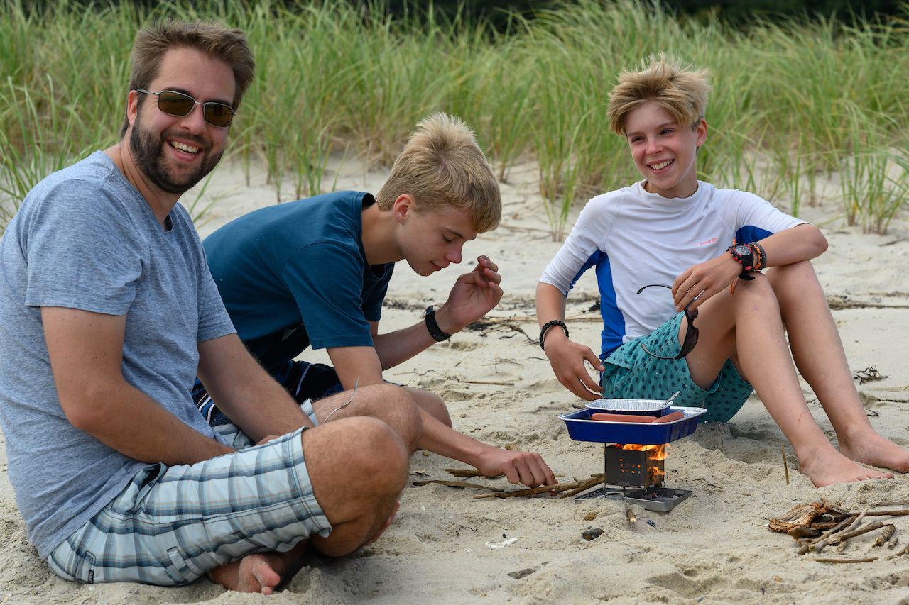 Three people sit on the beach, cooking hot dogs over a small portable grill while smiling and enjoying the moment.