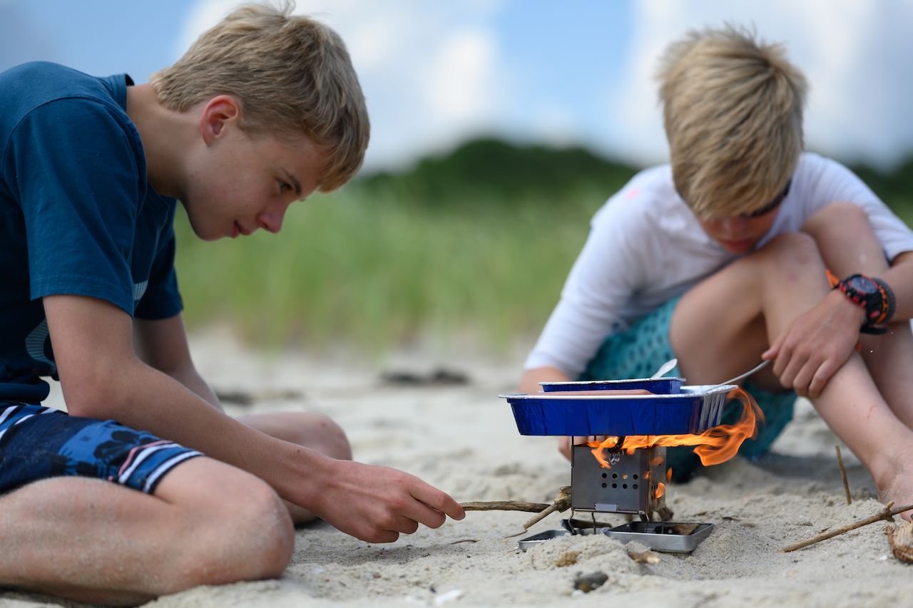 Two boys cook hot dogs over a small portable stove on a sandy beach, using sticks to tend the fire.