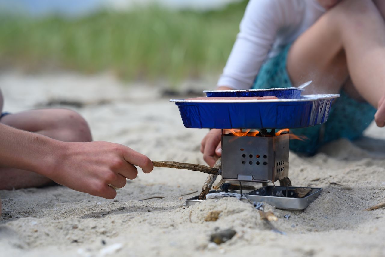 Two people cook hot dogs in foil trays over a small portable stove on a sandy beach.