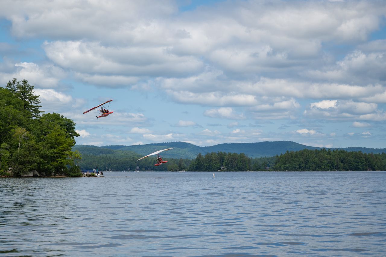 Two red ultralight seaplanes fly low over a calm lake, with green trees and mountains in the background.