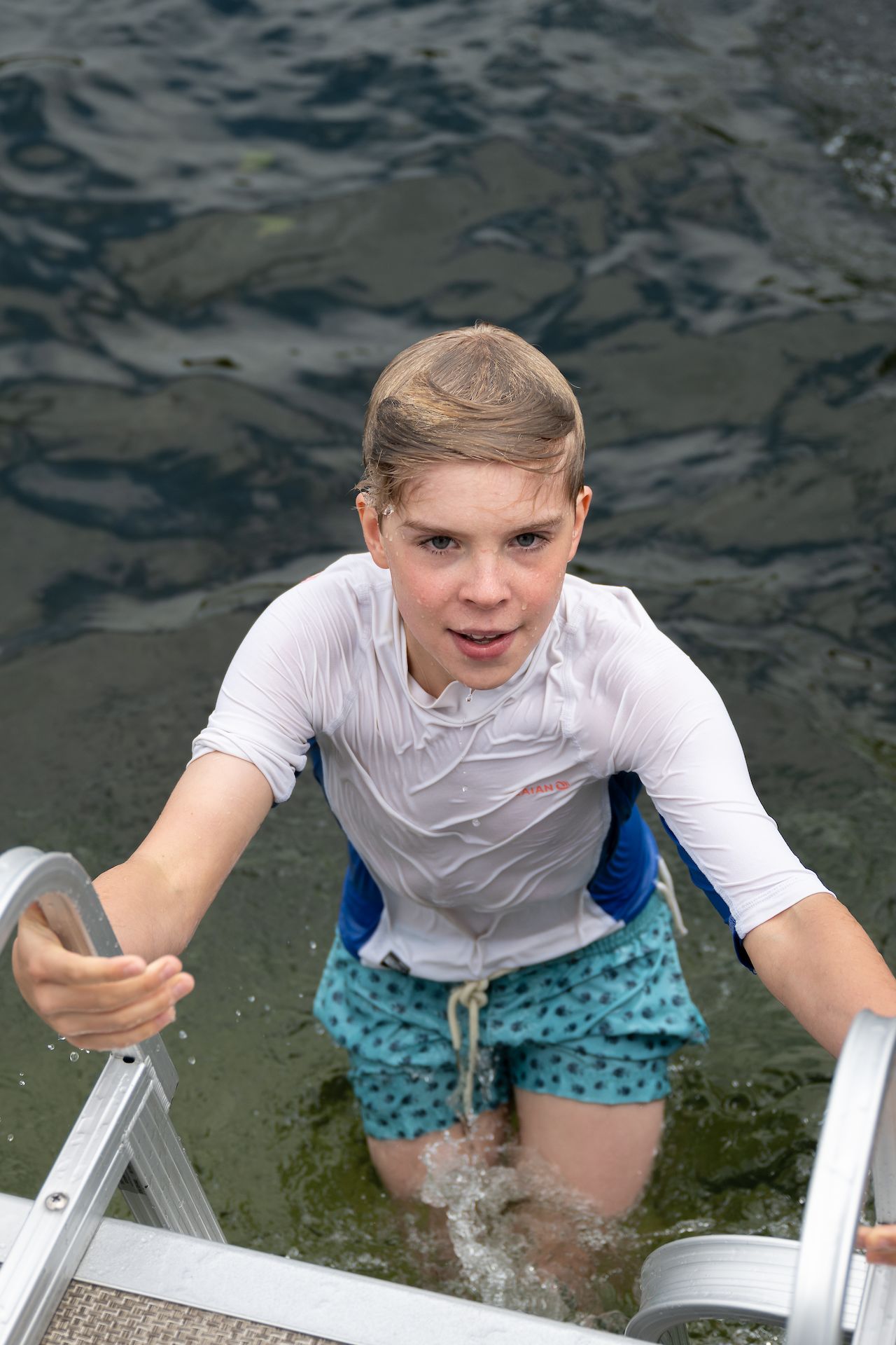 A person in a wet shirt and swim shorts climbs a metal ladder out of the lake.