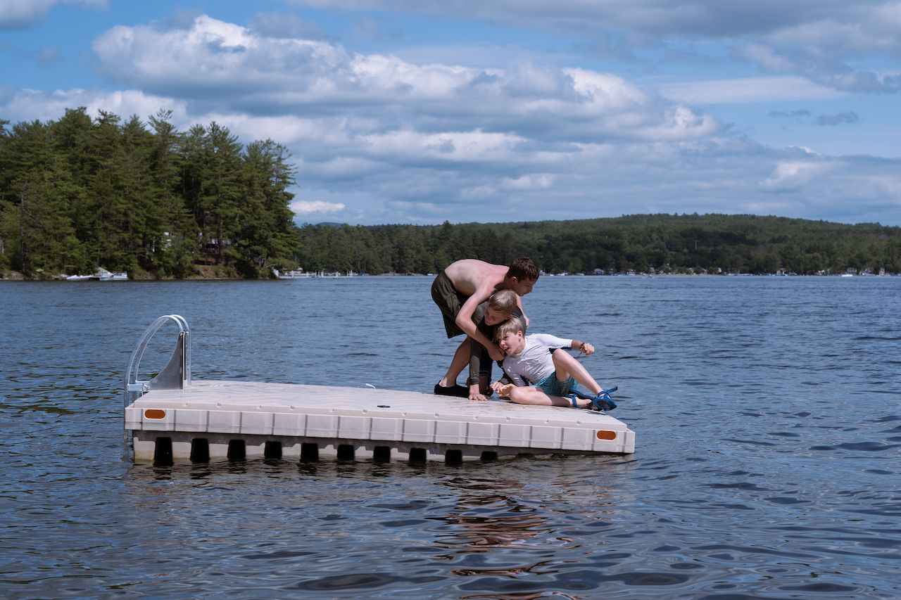 Three boys playfully wrestle on a floating dock, with one trying to pull another into the water.