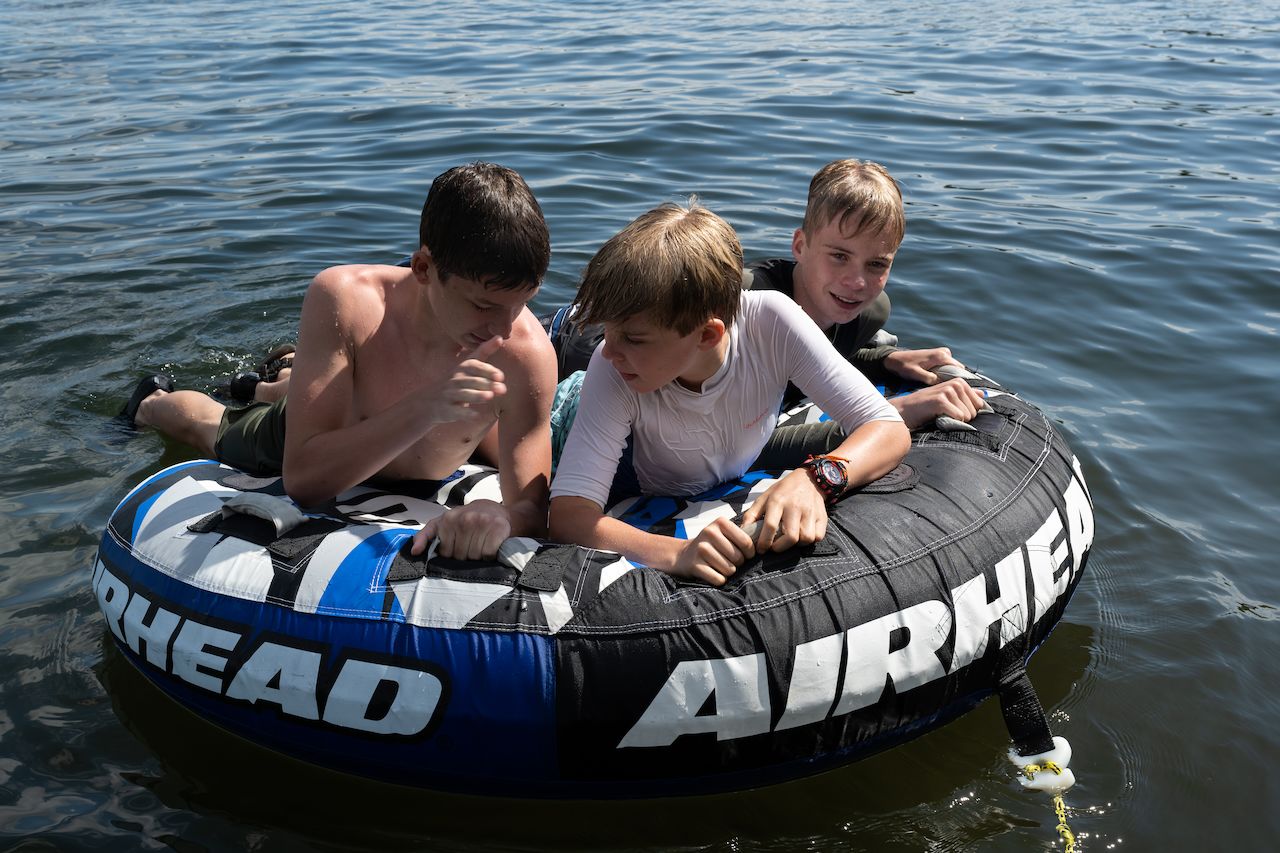 Three boys lay on a large inflatable tube floating on a lake, holding on and chatting.