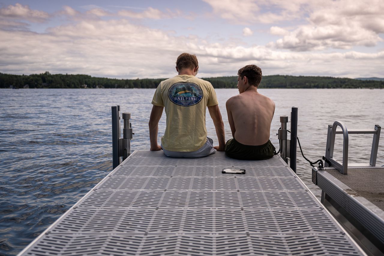Two people sit on a dock by the lake, facing the water.