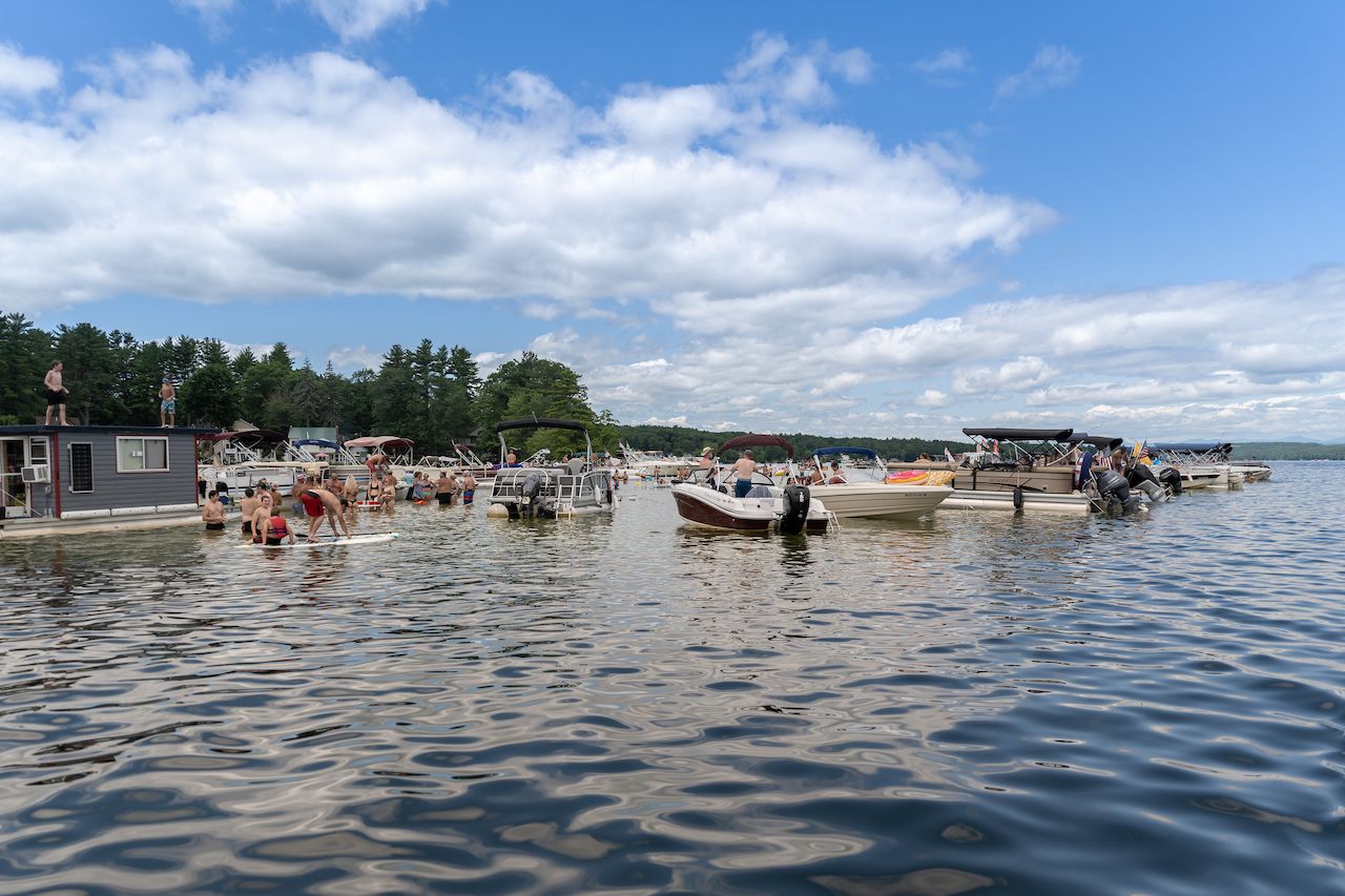 People wade in shallow lake water near anchored boats, with some standing on paddleboards and others on a floating house.
