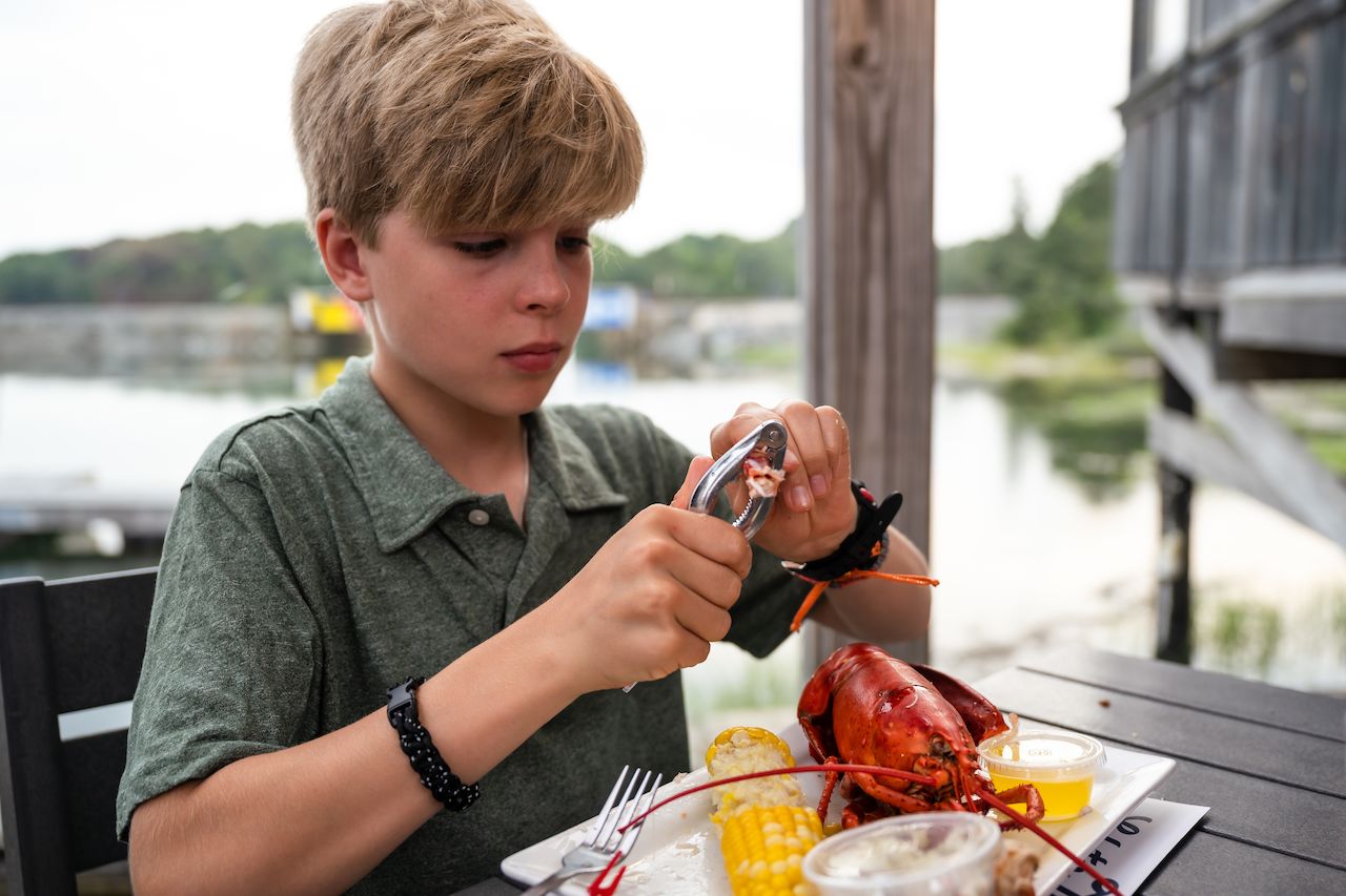 A young boy cracks open a lobster claw with a seafood cracker while eating at an outdoor table.