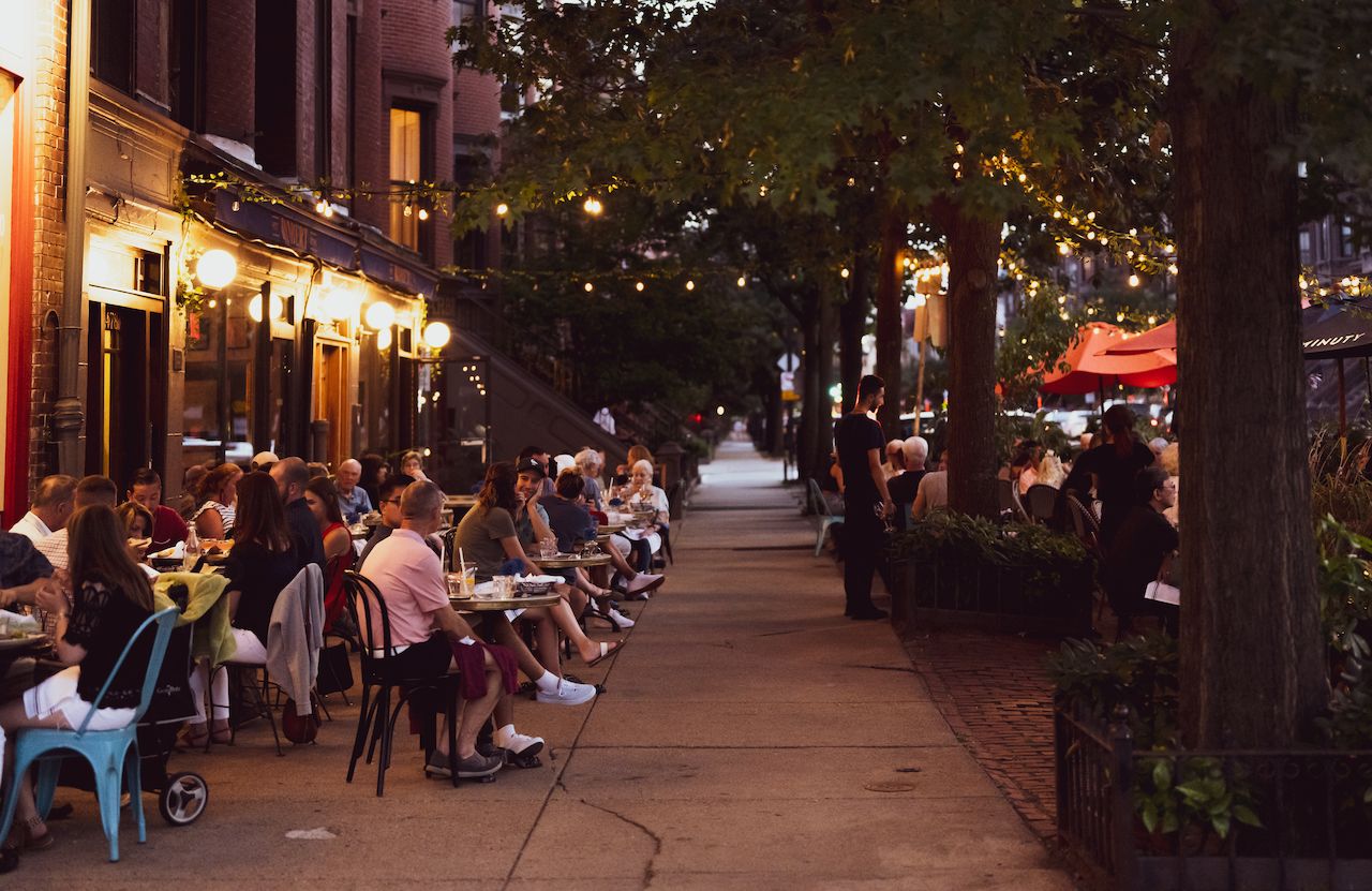 People dining at outdoor tables along a city sidewalk in the evening, with string lights and a waiter nearby.