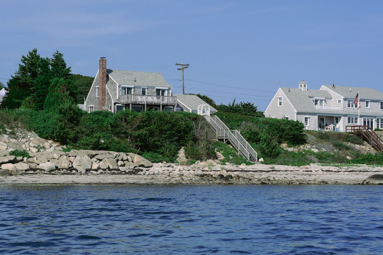 A gray rental house with a balcony and stairs leading to the rocky shoreline by the water.