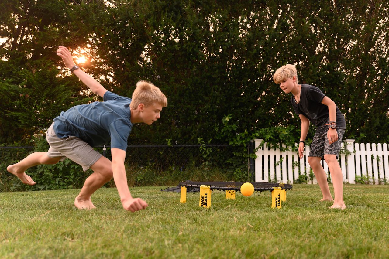 Two barefoot teenagers play Spikeball on grass, one lunging forward to hit the ball while the other watches.