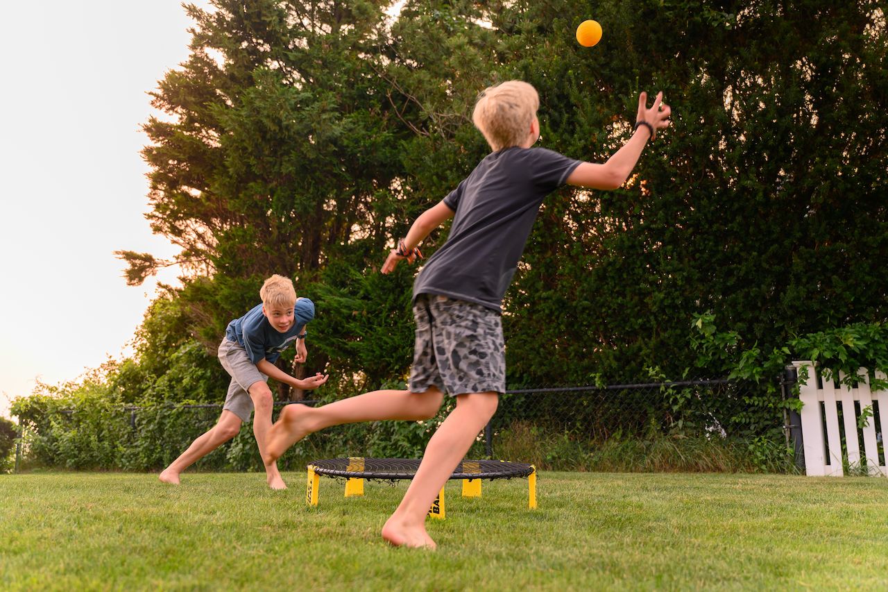 Two boys play Spikeball on a grassy field, one reaching to hit the ball while the other prepares to react.