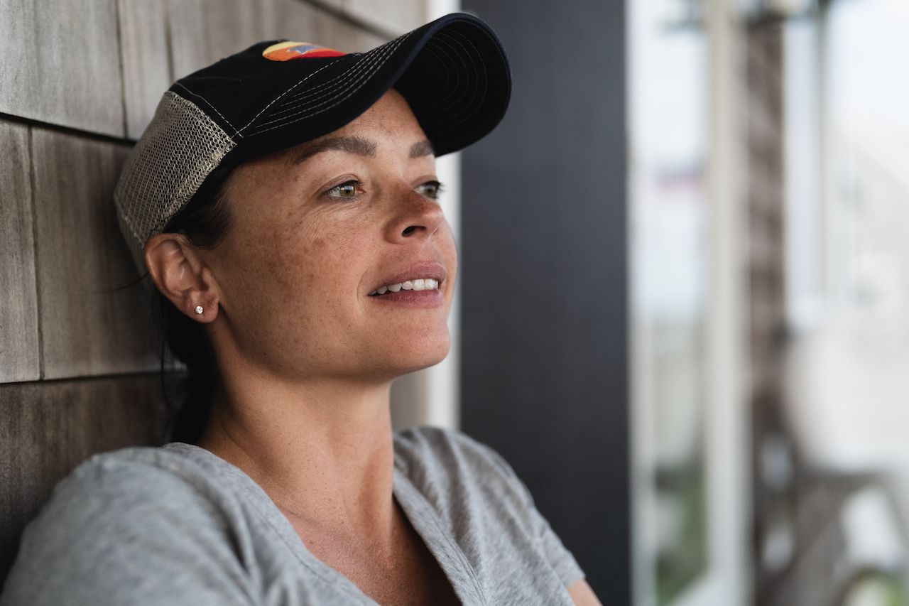 A woman in a cap leans against a wall, gazing into the distance with a relaxed expression.