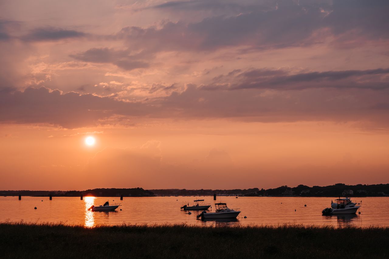 The sun sets over a calm bay with several boats floating on the water.