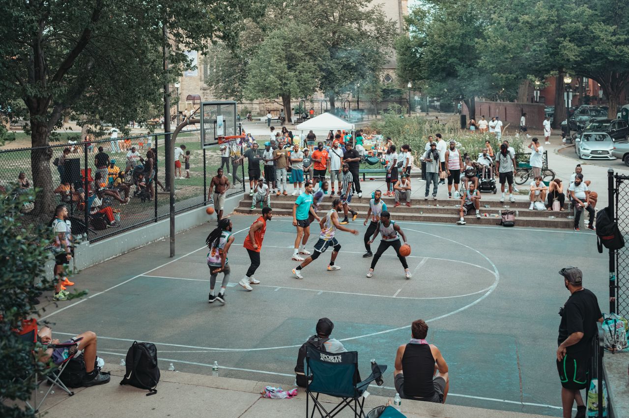 Players compete in a basketball game at an outdoor court, with spectators watching from the sidelines and steps.