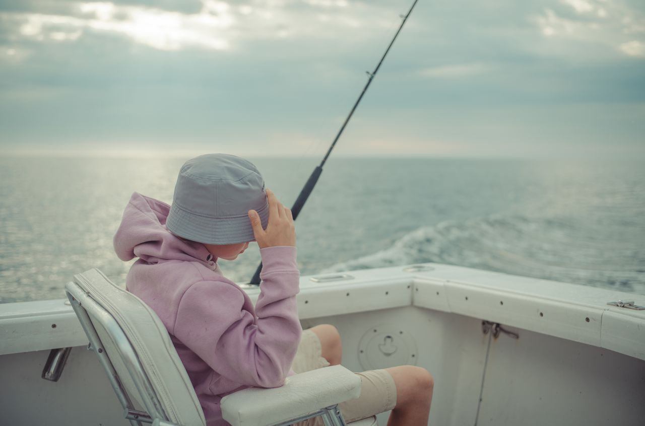 A person in a bucket hat and hoodie sits on a boat, holding a fishing rod while looking at the water.