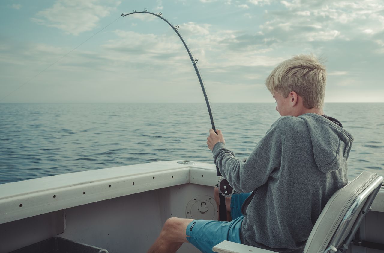 A person sitting on a boat, reeling in a fish with a fishing rod over the water.