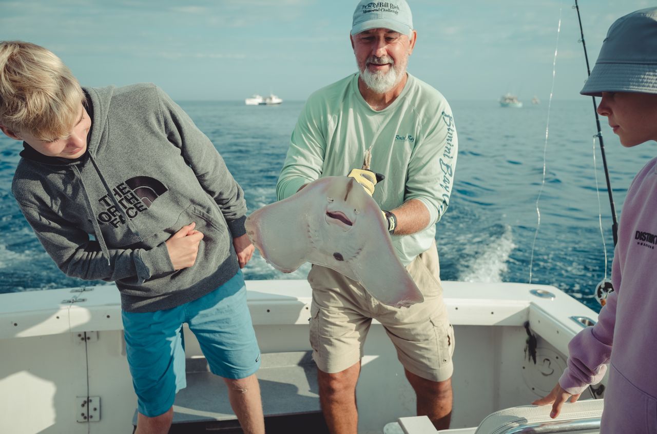 A man on a boat holds up a large skate while two others look at it.