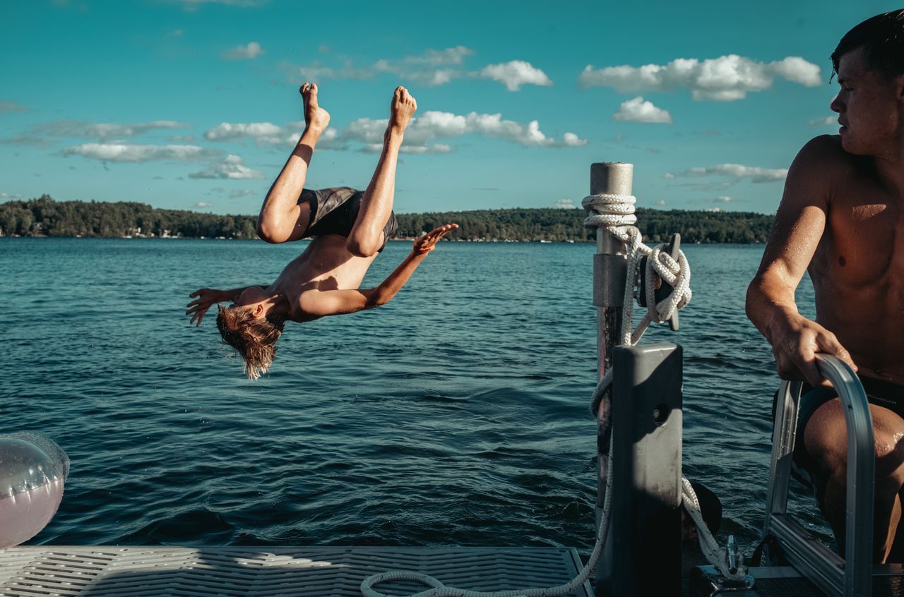 A person is mid-air doing a backflip off a jetty into the water, while another watches nearby.