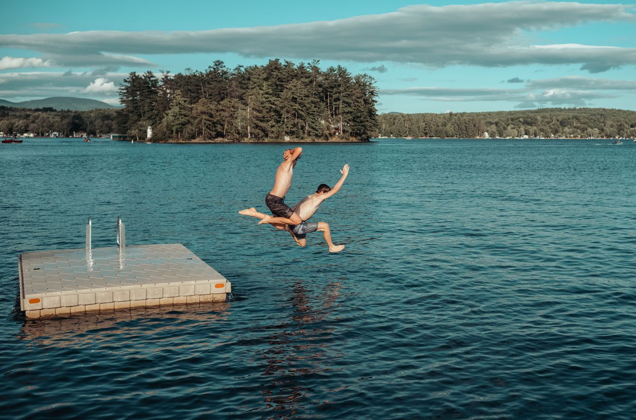 Two people jump off a floating dock into a lake, enjoying a summer swim in New Hampshire.