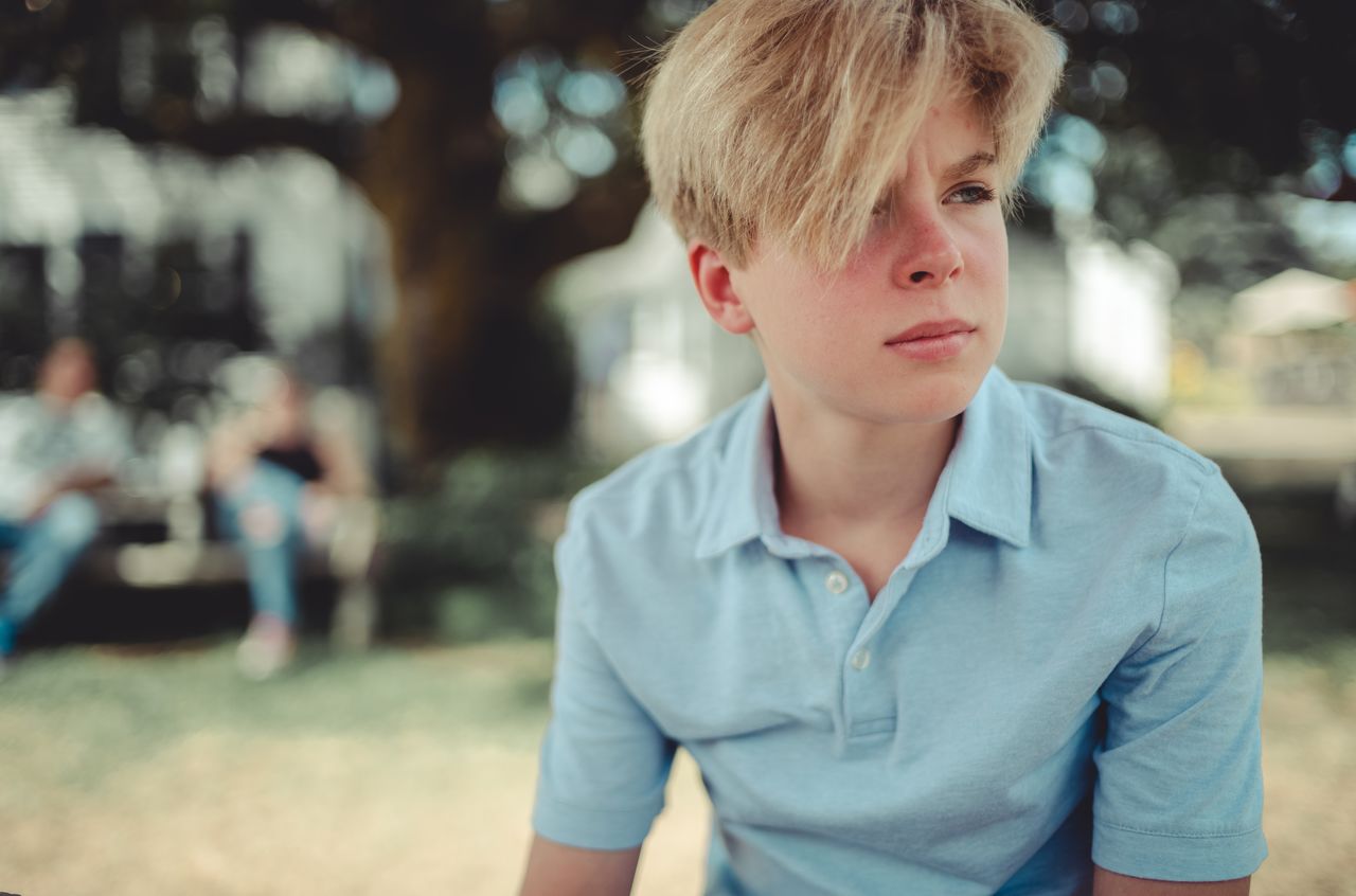 A young person in a light blue shirt sits outside, looking off into the distance with a thoughtful expression.