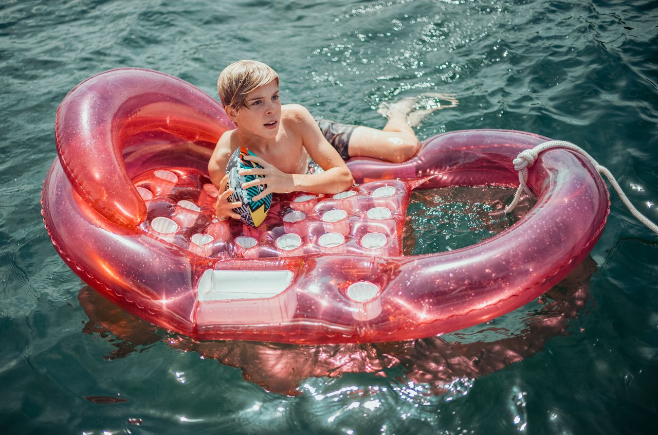 A boy is lying on a pink inflatable float in the lake, holding a small football.