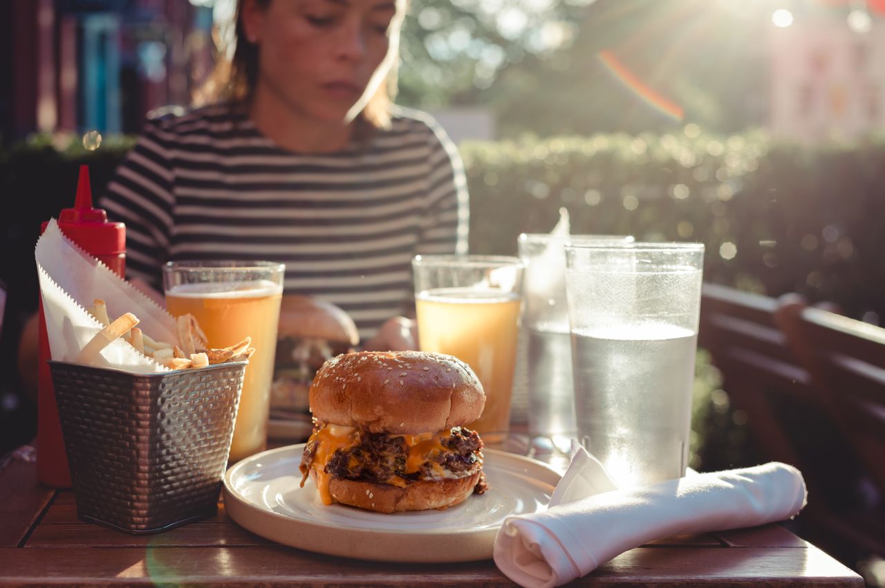 A cheeseburger with melted cheese sits on a plate at an outdoor table, with fries and drinks nearby.