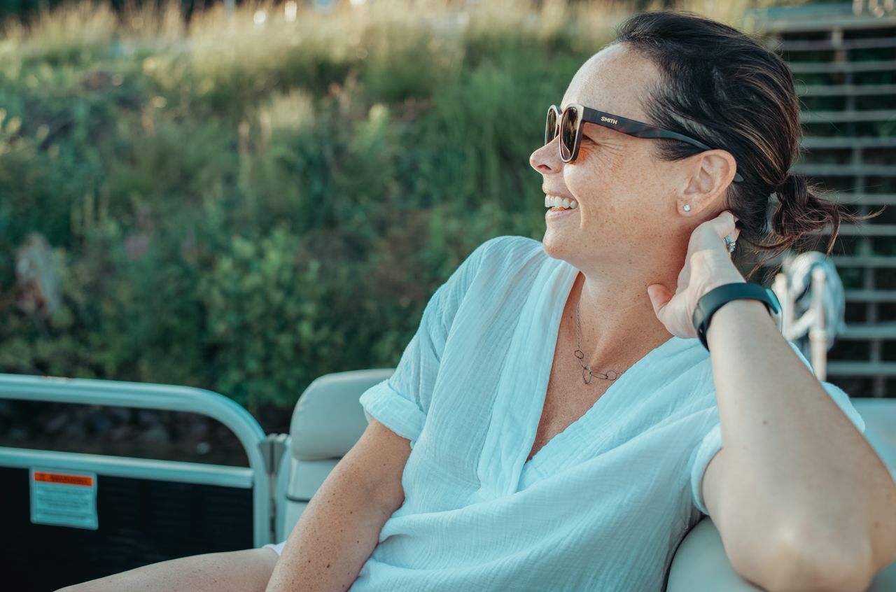 A woman wearing sunglasses and a white shirt smiles while relaxing on a boat.