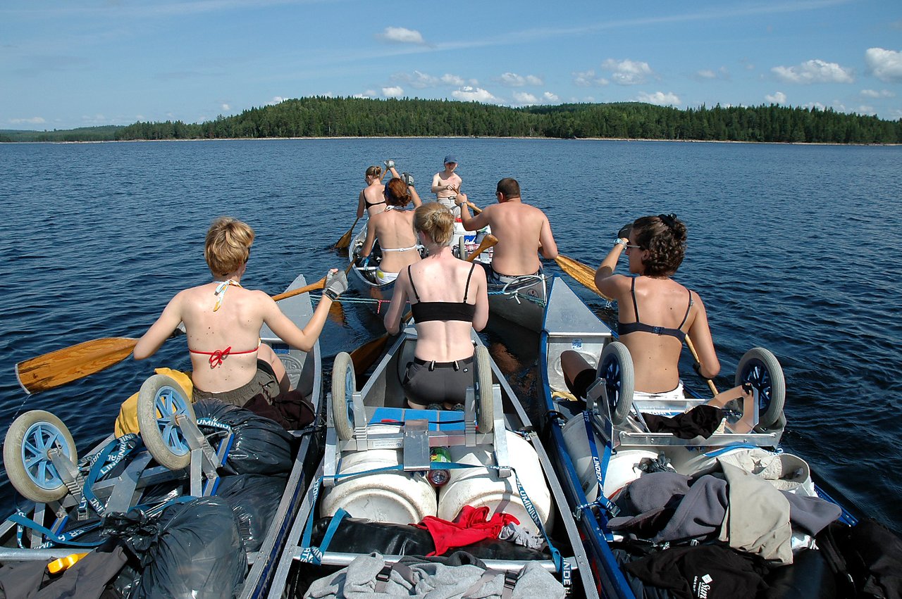 A group of people paddles canoes tied together on a lake, carrying gear and supplies under a clear sky.