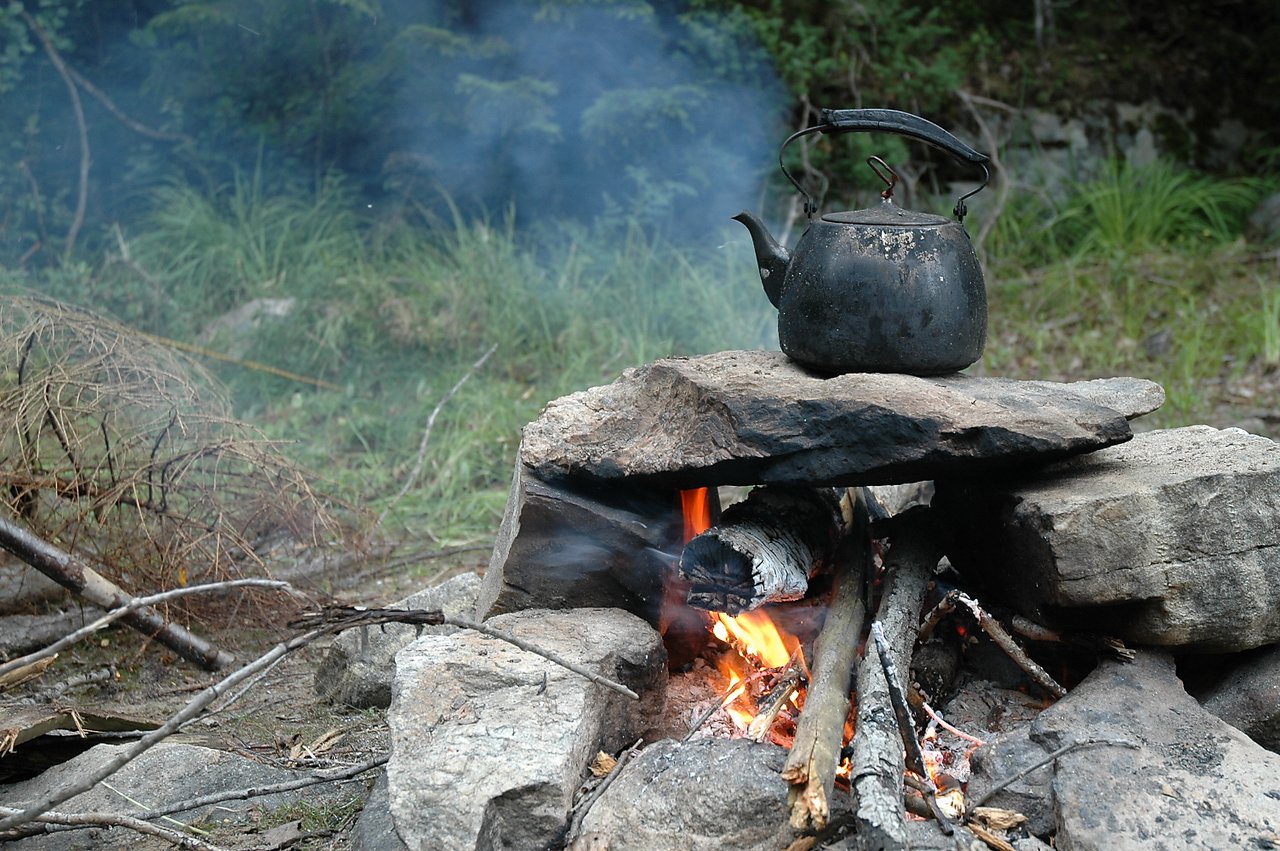 A black kettle sits on a stone fire pit, boiling water over an open flame in an outdoor setting.