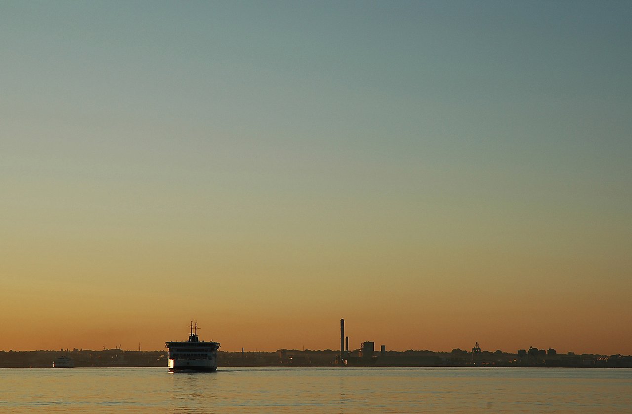 A ferry sails across calm water toward the shore, with a city skyline in the background at sunset.