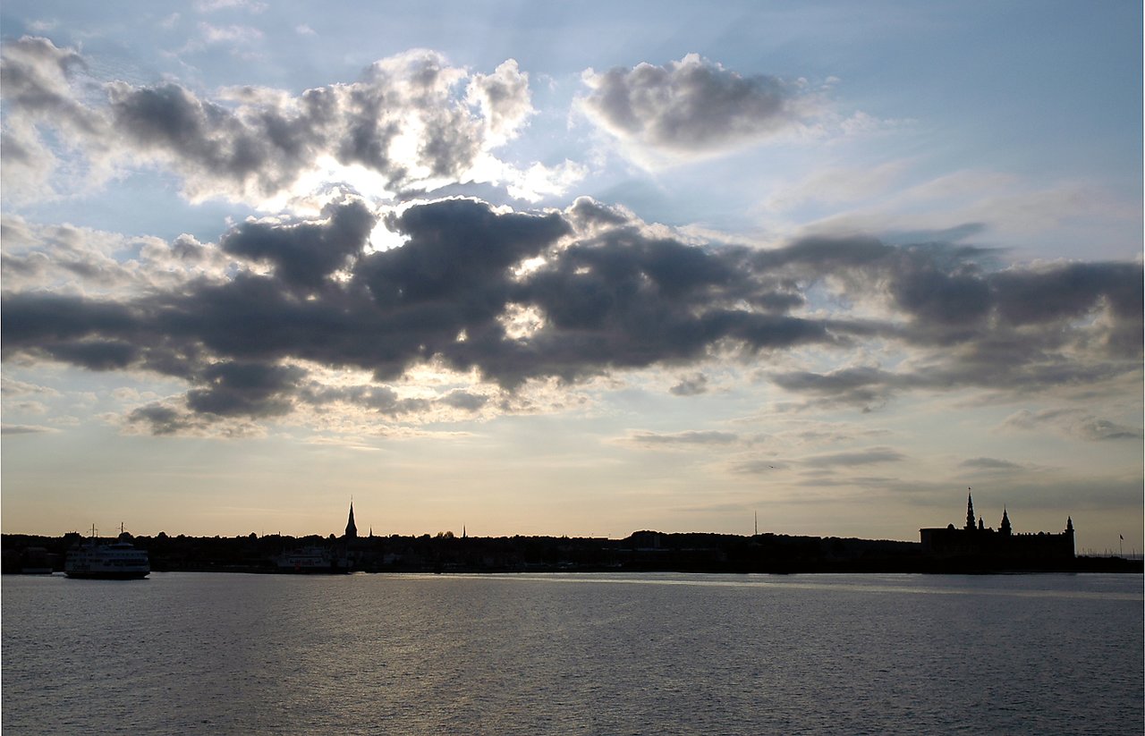 A waterfront view of Denmark at sunset, with a silhouetted cityscape, a castle, and a ferry on the water.