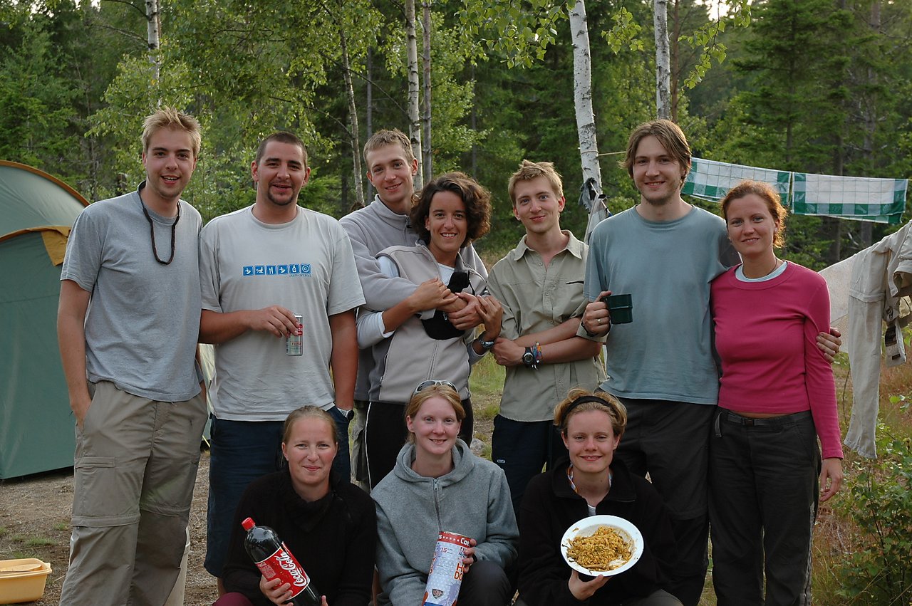 A group of ten people posing outdoors, some holding food and drinks, with tents and trees in the background.
