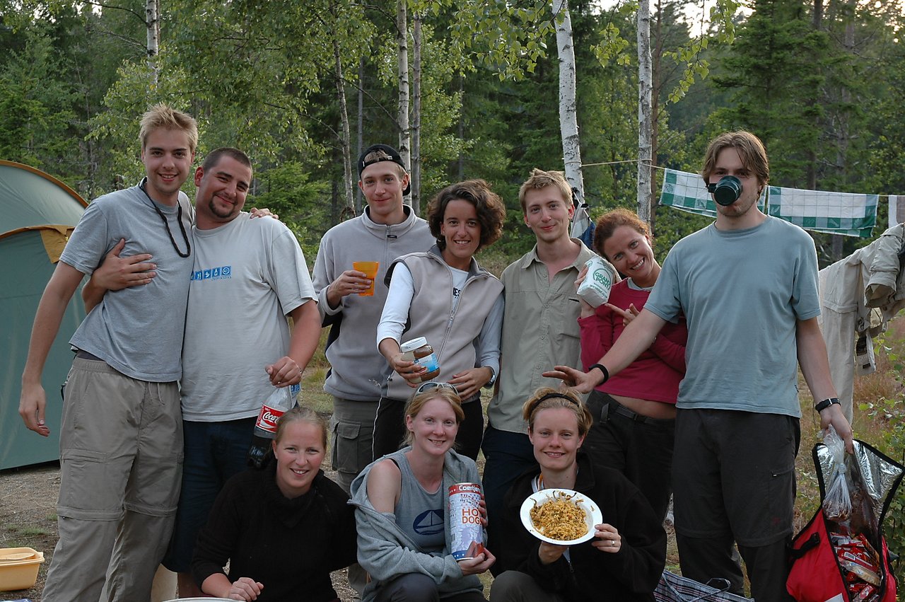 A group of friends posing at a campsite, holding food and drinks, smiling and enjoying their time together outdoors.